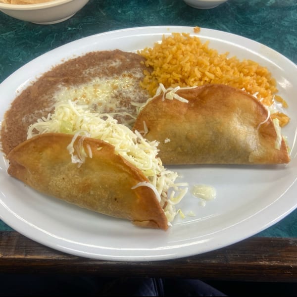 Two crispy tacos with shredded cheese, refried beans, and Mexican rice on a white plate.