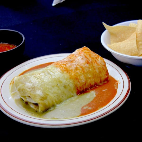 A large burrito covered in green and red sauce, with a side of tortilla chips and salsa.