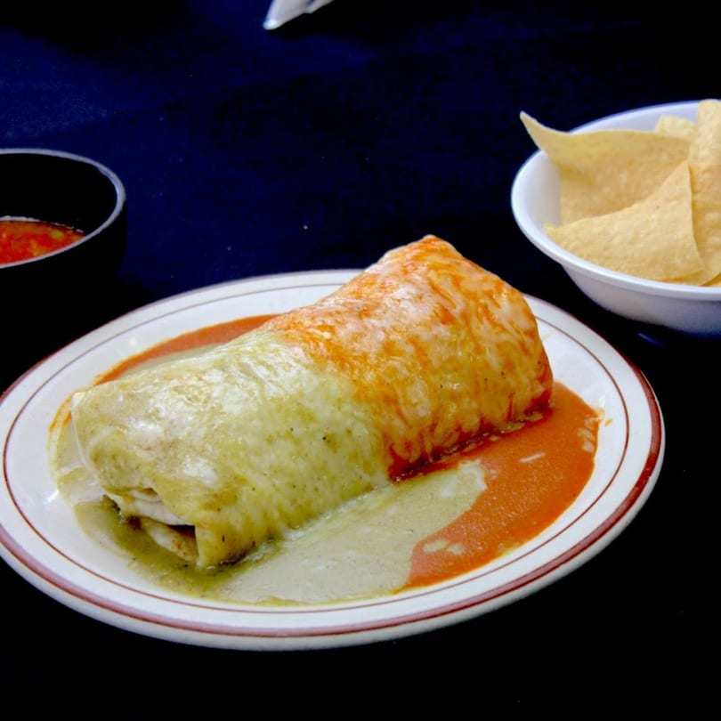 A large burrito covered in green and red sauce, with a side of tortilla chips and salsa.