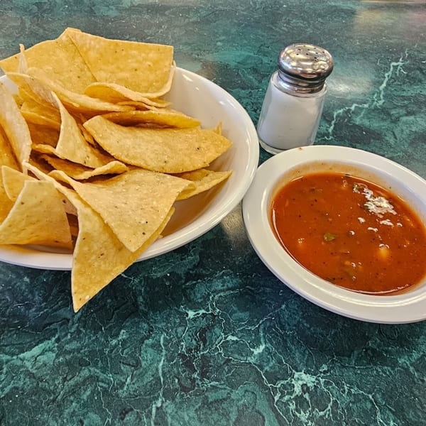 A bowl of tortilla chips, a dish of salsa, and a salt shaker on a green marbled table.