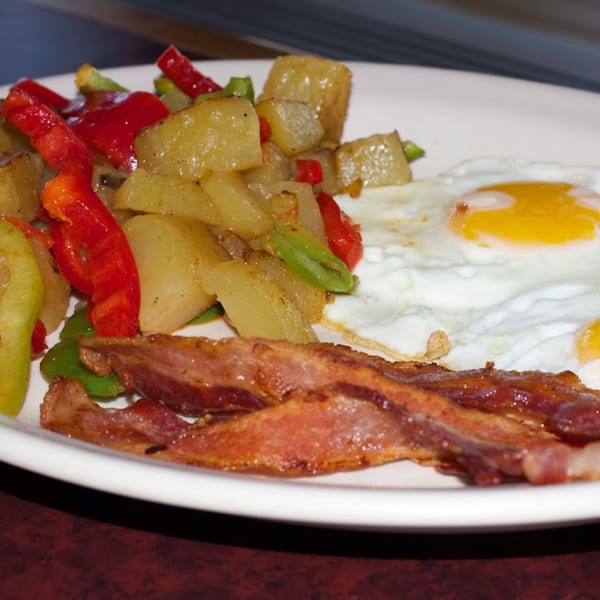 A breakfast plate with crispy bacon, fried eggs, and seasoned potatoes with red and green peppers.